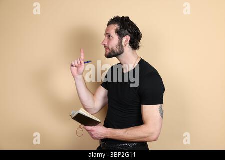 Un jeune homme dans un t-shirt noir et un pantalon tient un cahier et un stylo, regardant vers le haut et de côté avec son index levé et la bouche ouverte, comme s'il avait un su Banque D'Images