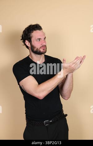 Un jeune homme portant un t-shirt noir et un pantalon tape des mains avec une expression heureuse ou pleine d'espoir tout en regardant vers le haut. Tatouage visible. Portrait studio o Banque D'Images