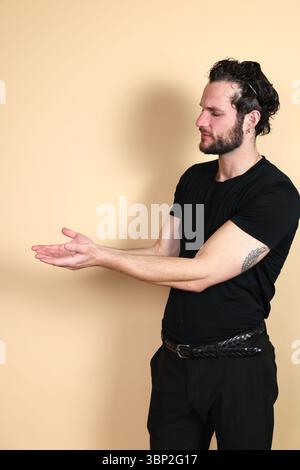 Un jeune homme dans un t-shirt noir et un pantalon, vu de profil latéral, regarde sur le côté et présente avec les deux paumes ouvertes vers le haut, comme s'il offrait ou Banque D'Images