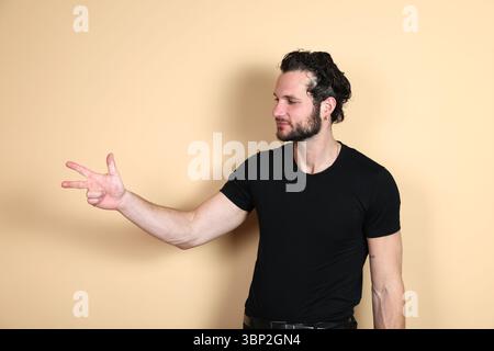 Un jeune homme portant un t-shirt noir tient trois doigts sur sa main, regardant vers sa main ou de côté, comme s'il comptait ou indiquait le chiffre trois. S Banque D'Images