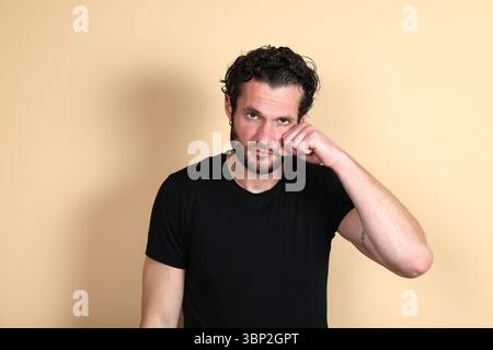 Un jeune homme portant un t-shirt noir regarde la caméra avec une expression fatiguée ou frustrée, frottant son œil avec son poing, la tête légèrement inclinée. Studio Banque D'Images