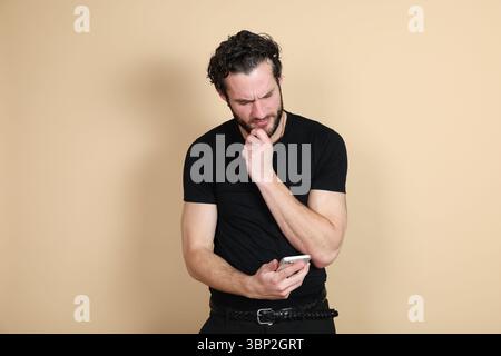 Un jeune homme portant un t-shirt noir et un pantalon regarde son smartphone avec une expression confuse ou réfléchie, la main sur son menton. Portrait de studio Banque D'Images