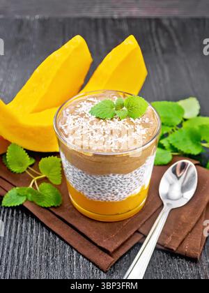 Dessert en couches de purée de citrouille, yaourt, cacao et graines de chia dans un verre, tranches de légumes, cuillère et menthe sur une serviette sur fond de planche de bois Banque D'Images