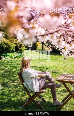 Femme assise dans le jardin de printemps sous le cerisier en fleurs et profitant de la journée ensoleillée. Détente en plein air Banque D'Images