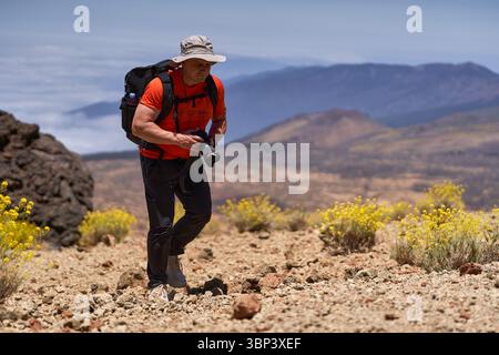 Homme randonnée à travers un terrain volcanique rocheux avec caméra à la main et paysage de montagne en arrière-plan Banque D'Images