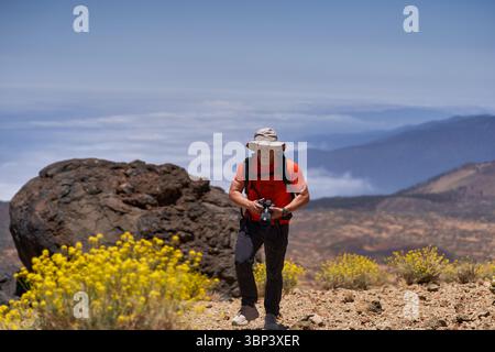 Homme randonnée à travers un terrain volcanique rocheux avec caméra à la main et paysage de montagne en arrière-plan Banque D'Images