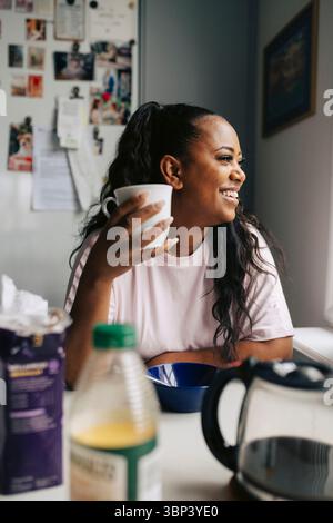 Femme heureuse tenant une tasse regardant loin tout en étant assis à table dans la maison Banque D'Images