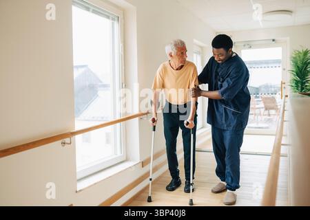 Soignant masculin aidant un homme âgé marchant avec des béquilles dans le couloir d'une maison de retraite Banque D'Images