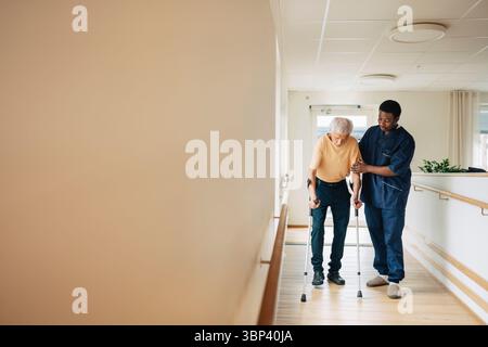Thérapeute masculin aidant l'homme âgé marchant avec des béquilles dans le couloir à la maison de retraite Banque D'Images