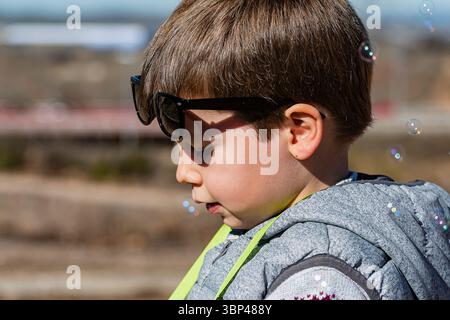 Un enfant aime souffler des bulles tout en portant des lunettes de soleil ludiques dans un endroit extérieur ensoleillé. Banque D'Images
