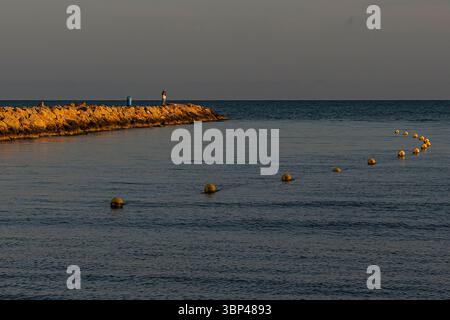 Un individu se tient sur une jetée rocheuse au coucher du soleil, contemplant la mer calme avec des bouées flottantes en vue. Banque D'Images
