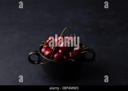 Cerises fraîches mûres juteuses dans une assiette noire sur fond noir. Fruits de saison, récolte. Mise au point sélective, gros plan. Banque D'Images