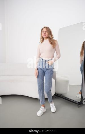 Une femme dans un pull léger et un Jean bleu se tient sur un sol gris, main dans la poche, près d'un canapé blanc ondulé et d'un miroir debout reflétant son image Banque D'Images