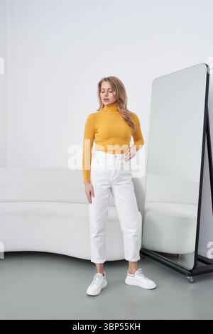 Femme se tient en toute confiance dans une tenue élégante, avec un col roulé jaune et un pantalon blanc. Elle pose à côté d'un grand miroir, contre une inte minimaliste Banque D'Images