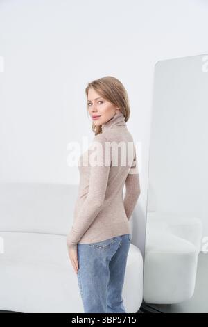 Jeune femme en décontracté col roulé beige et Jean bleu pose en toute confiance près d'un grand miroir, canapé visible. Élégant, avec une expression douce et détendue Banque D'Images