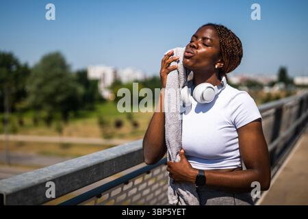 Portrait de femme africaine fatiguée dans des vêtements de sport essuyant la sueur de son visage avec une serviette et se reposant après un entraînement sportif intense sur le pont de la ville. Banque D'Images