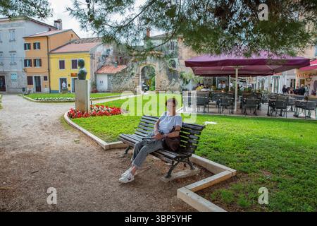 Porec, Croatie - 21 mai 2025 : femme caucasienne d'âge moyen se relaxant sur un banc dans un petit parc entouré de bâtiments historiques et d'un café en plein air Banque D'Images