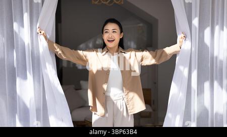 La femme asiatique excitée ouvrant des rideaux blancs laisse la lumière du soleil entrer dans la pièce Banque D'Images