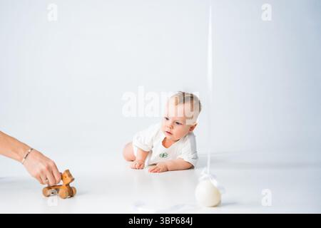 Bébé curieux rampant sur le sol vers le jouet en bois sur scène de studio de fond blanc. Banque D'Images