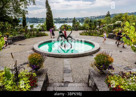 Les visiteurs se promènent dans les magnifiques jardins de l'île aux fleurs de Mainau à Constance, en Allemagne. Au centre de l'arène des fontaines se trouve une fontaine ronde ornée d'une sculpture moderne, entourée de parterres de fleurs colorées et d'une végétation luxuriante. Le lac de Constance peut être vu en arrière-plan avec le paysage riverain en face, créant une atmosphère idyllique Banque D'Images