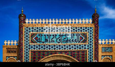 Une vue rapprochée d'une arche carrelée colorée présentant des motifs géométriques complexes et une calligraphie arabe, placée sur un ciel bleu vif. Banque D'Images