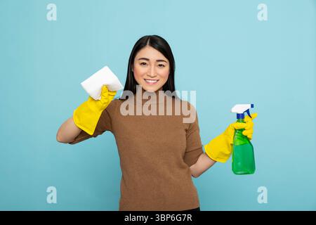 Femme au foyer asiatique prête à l'action, tenant une bouteille de détergent et une serviette en papier avec un sourire joyeux. Le nettoyant féminin heureux dans les gants en caoutchouc saisit les produits de nettoyage, désireux de commencer les tâches ménagères. Banque D'Images