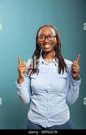Femme afro-américaine souriante pointe vers le haut avec les deux index, soulignant l'espace pour le contenu promotionnel. Modèle féminin noir en chemise bleue, regardant la caméra et faisant des gestes ci-dessus. Banque D'Images
