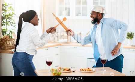 Drôle de couple de Black ayant l'amusement dans la cuisine, en utilisant des ustensiles de cuisine pour jouer combat Banque D'Images