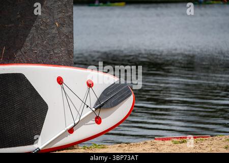 Gros plan d'une planche de paddleboard avec une pagaie sur une plage de sable à côté d'un plan d'eau. L'accent est mis sur l'équipement de sports nautiques. Banque D'Images