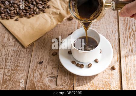 Café frais breveté dans cezve (cafetière turque traditionnelle) la main de la femme verse le café dans une tasse blanche Banque D'Images