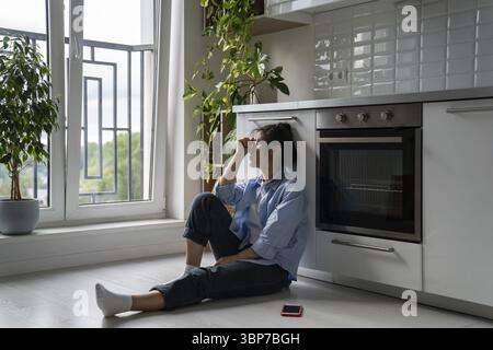 Malheureuse fille asiatique assise sur le sol dans la cuisine se sentant déprimée après l'utilisation des médias sociaux, étant ignorée par petit ami, traitant du rejet dans l'amour. Banque D'Images