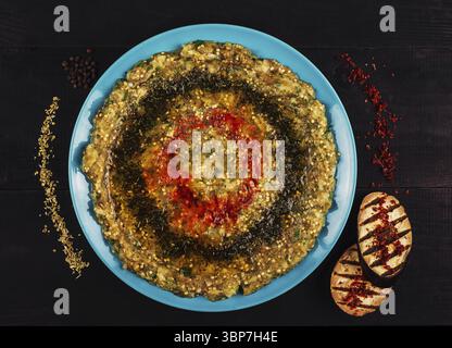 Nourriture, Baba ghanoush décoré d'huile d'olive et d'épices est couché sur une plaque bleue à côté de tranches d'aubergines grillées et d'épices dispersées sur un bois noir Banque D'Images