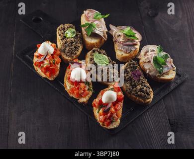 Nourriture, délicieuse bruschetta avec tomates, mozzarella, pâté de viande et hareng couché sur une planche à découper en pierre noire Banque D'Images