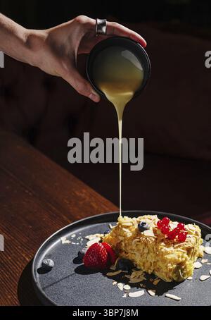 Nourriture, Une main verse élégamment une sauce dorée sur une tranche de gâteau ornée de baies fraîches et d'amandes sur une assiette sombre. L'ambiance chaleureuse rehausse la Banque D'Images