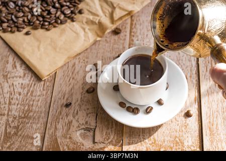 Café frais breveté dans cezve (cafetière turque traditionnelle) la main de la femme verse le café dans une tasse blanche Banque D'Images