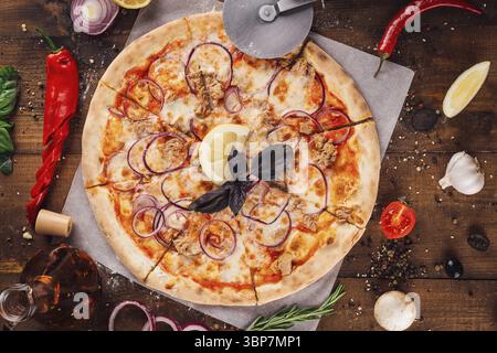 Nourriture, pizza au thon fraîchement cuit avec des rondelles d'oignon rouge et une tranche de citron couché sur une table en bois entourée d'ingrédients frais Banque D'Images