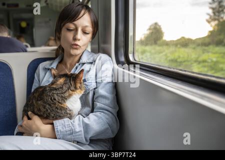 Fille satisfaite embrassant le chat sur les baisers soufflant les genoux, exprimant l'affection de l'amour. Propriétaire femme câlin avec Kitty détente se reposant dans le siège dans le wagon. Jou Banque D'Images