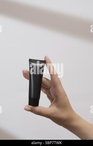 La main de la femme tient une maquette de tube noir de crème. Emballage non étiqueté pour les cosmétiques à la lumière du soleil sur fond blanc. Concept de soin de la peau. Alose dure Banque D'Images