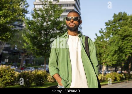 Réfléchi jeune homme afro-américain dans des lunettes de soleil va sur le trottoir confiant regarde devant. Sérieux élégant gars noir étudiant dans des lunettes de soleil marche dans c Banque D'Images