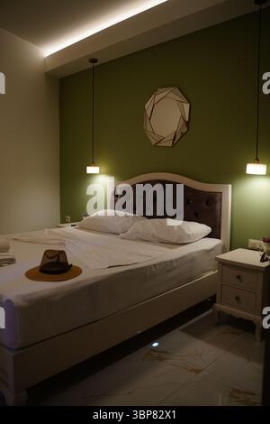 Chambre élégante avec un mur vert, une tête de lit touffetée et des suspensions modernes. Un chapeau de paille repose sur le lit fait, ajoutant une touche de confort et Banque D'Images