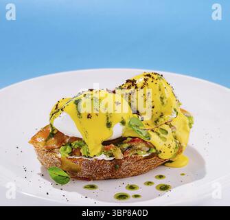 Nourriture, deux œufs parfaitement pochés assis sur le pain grillé avec avocat et saumon, arrosé d'une sauce jaune crémeuse et entouré d'herbes fraîches Banque D'Images