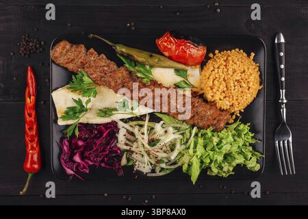 Nourriture, traditionnel turc adana kebab servi sur une assiette avec tomate grillée, poivre, oignon, salade, persil, pain lavash et riz pilaf bulgur sur bla Banque D'Images