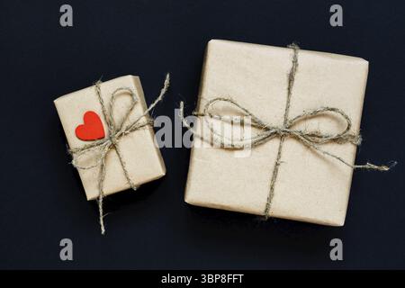 Boîte-cadeau fabriquée à la main enveloppée de papier artisanal avec coeur en bois rouge, corde et noeud sur fond noir. Vue de dessus, plan d'appartement Banque D'Images