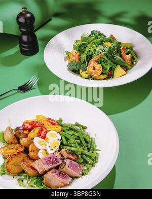 Nourriture, deux assiettes blanches avec de savoureuses salades fraîches avec des crevettes, thon, roquette, tomates et œufs sur fond vert Banque D'Images