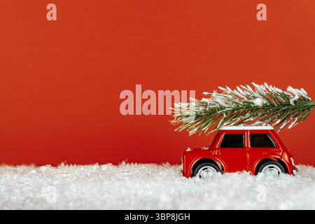 Petite voiture de jouet rouge transportant un arbre de spruse sur une route enneigée. Carte de vœux de Noël festif Banque D'Images