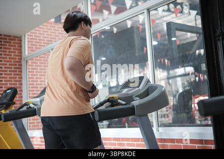 Un homme court sur un tapis roulant dans une salle de gym. Il porte une chemise orange et il est concentré sur son entraînement Banque D'Images