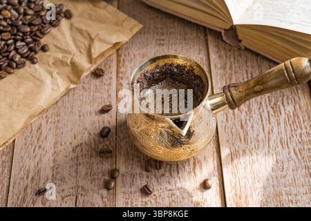 Café frais breveté dans cezve (cafetière turque traditionnelle) livre ouvert et grains de café sur la table en bois Banque D'Images