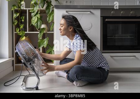 Surchauffe à la maison. Joyeuse jeune femme asiatique assise devant un ventilateur de sol électrique à la maison, rafraîchissant pendant les chaudes journées d'été Banque D'Images