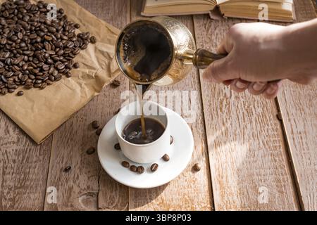 Café frais breveté dans cezve (cafetière turque traditionnelle) la main de la femme verse le café dans une tasse blanche Banque D'Images