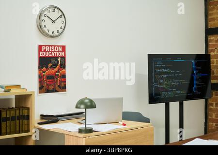 Scène de salle de classe vide montrant un bureau en bois avec ordinateur portable ouvert, des papiers éparpillés, une lampe de bureau verte, une étagère avec des livres de droit, une horloge murale, une affiche d'éducation en prison, un grand moniteur affichant le code de programmation Banque D'Images